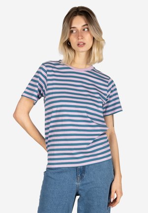 Junge Frau mit kurzen blonden Haaren, die ein pink-grün gestreiftes T-Shirt und blaue Jeans trägt und seitlich blickt, mit einer Hand auf der Hüfte.