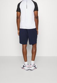 Miestens sportinė apranga, susidedanti iš baltos trumpomis rankovėmis marškinėlių su juodais raštais ant rankovių ir tamsiai mėlynų šortų, derinama su baltais sportiniais bateliais.