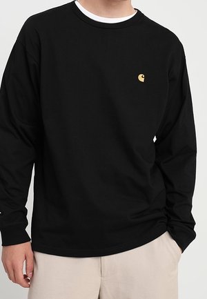 Homme portant un t-shirt Carhartt noir à manches longues avec un petit logo jaune sur la poitrine et un pantalon beige, debout avec une main dans la poche.