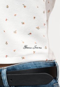 Λευκή μπλούζα με μικρά πορτοκαλί ανθισμένα σχέδια και κέντημα με τη φράση "Guess Jeans", συνδυασμένη με μπλε τζιν παντελόνι και μαύρη ζώνη στη μέση.