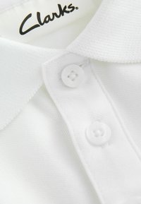 Närbild av en vit polo t-shirt med krage och två vita knappar, där "Clarks"-lappen är synlig inuti kragen.