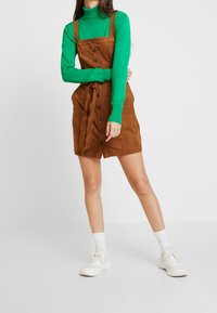 Braunes Wildlederkleid mit Knöpfen an der Vorderseite, einem Bindegürtel und Schulterträgern, kombiniert mit einem grünen Rollkragenpullover und weißen Sneakers.