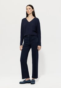 Junge Frau steht und trägt einen dunkelblauen V-Ausschnitt-Pullover, passende weit geschnittene Jeans und Denim-Slipper vor einem schlichten weißen Hintergrund.