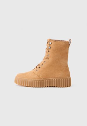 Botte en suède beige à lacets avec semelle en caoutchouc épaisse à rainures et tirette, sur fond uni, vue de côté.