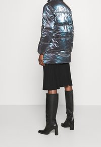 Iridescent pufferjacka med horisontella sömmar, svart plisserad kjol och knähöga svarta läderstövlar med klack.