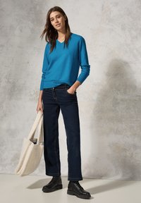 Maglione blu a V e jeans scuri con orlo arrotolato; accessorato con una borsa a tracolla crema e pelosa e stivali neri con lacci alla caviglia.