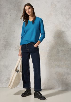 Blauer V-Ausschnitt-Pullover und dunkelblaue Jeans mit aufgerolltem Saum; accessoriert mit einer cremefarbenen, flauschigen Tote-Bag und schwarzen Schnürstiefeletten.