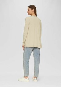 Beige sweatshirt met lange mouwen, een relaxte pasvorm, zijsplitten en een zachte textuur, gecombineerd met lichtblauwe jeans en witte sneakers.