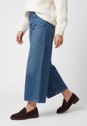 Person iført blå jeans med vide ben, hvid striktrøje og mørkebrune slip-on loafers, stående mod en ensfarvet lys baggrund.