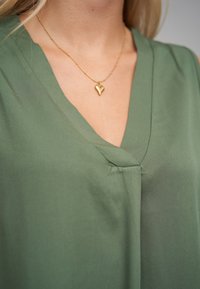 Grønn, lys stofftopp med V-utringning og en diskret glans. Et gyldent hjerteformet smykke med en tynn kjede komplimenterer utringningen.