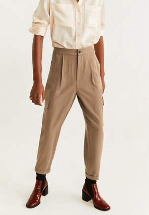 Persona con pantalones cargo beige con botón y bolsillos, blusa crema con mangas remangadas, calcetines negros y zapatos marrones de tacón.