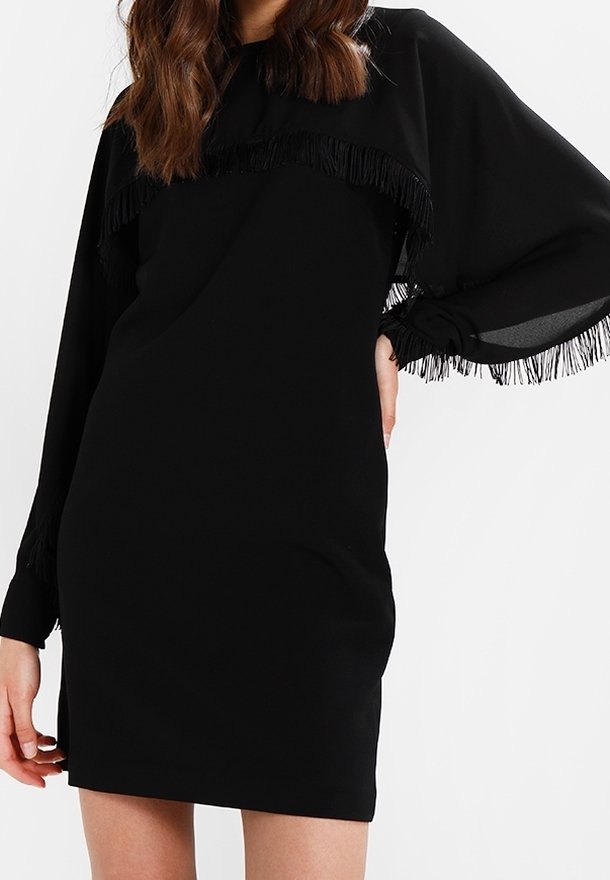 Femme portant une robe noire à manches longues avec des détails en franges sur la poitrine, les manches et les poignets, debout devant un fond uni.