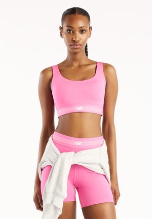 Femeie cu părul împletit, purtând un sutien sport roz și pantaloni scurți asortați, cu un hanorac alb legat în jurul taliei, stând pe un fundal alb.