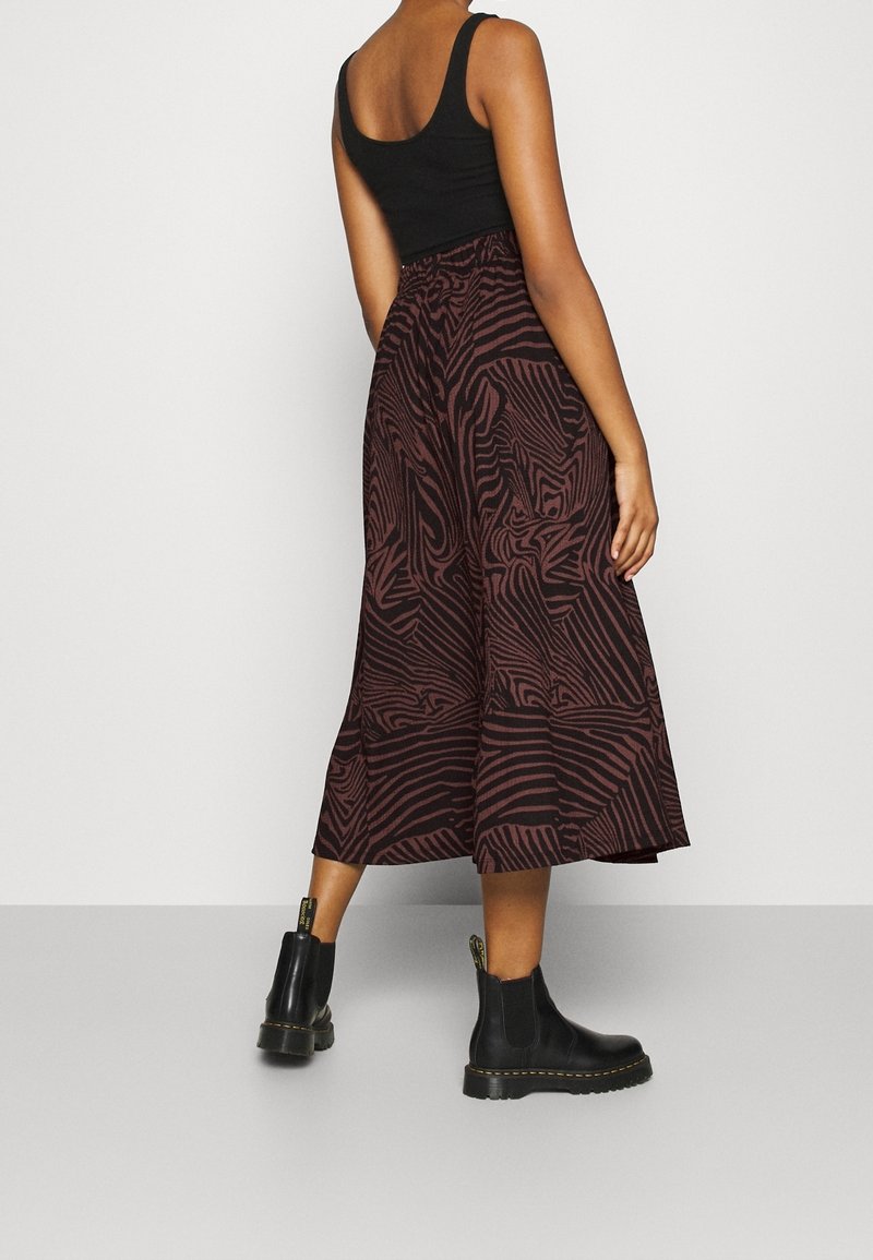 Femme portant un débardeur noir, un pantalon large à motif marron et noir, et des bottines noires, debout devant un fond uni.