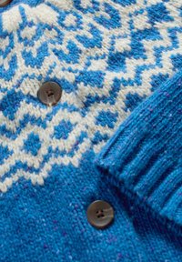 Neulepaita, jossa on sinivalkoinen kuvio geometrisine muotoineen, ribbireunukset ja tummanruskea nappidetaili. Pehmeä rakenne.