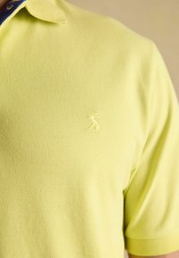 Limetková zelená polo košeľa z texturovanej tkaniny s malou vyšitou nášivkou na hrudi. Má klasický golier a krátke rukávy.