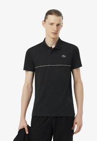Polo negro de algodón, con cuello abotonado, mangas cortas y una franja blanca contrastante en el pecho. Logo de Lacoste visible.