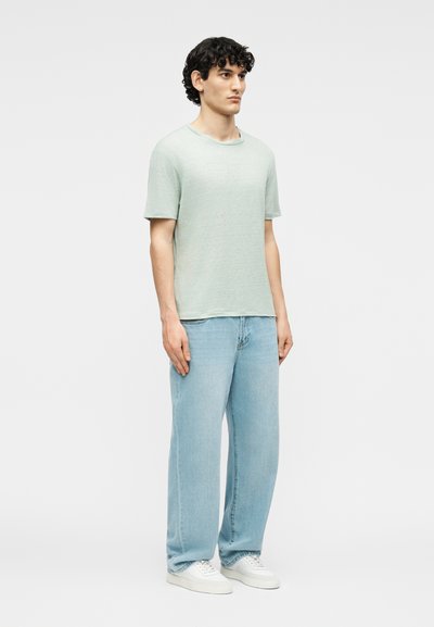 Νέος άνδρας που στέκεται με το σώμα ελαφρώς στραμμένο προς τα δεξιά, φοράει ανοιχτοπράσινο κοντομάνικο πουκάμισο, ανοιχτό μπλε φαρδιά τζιν και άσπρα αθλητικά παπούτσια.