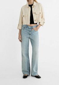 Světlounce béžová zkrácená bunda se zlatými knoflíky, v kombinaci s vysokým pasem, světle modrými širokými džíny a černými balerínkami. Hladká texturovaná látka.