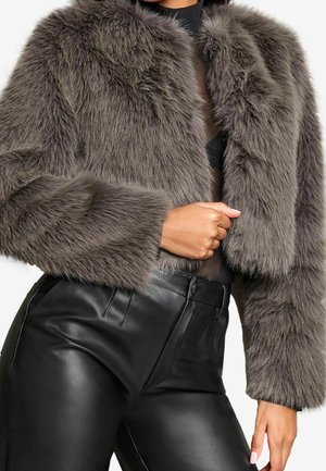 Femme portant une veste courte grise en fausse fourrure et un pantalon en cuir noir taille haute, une main reposant près de sa taille.