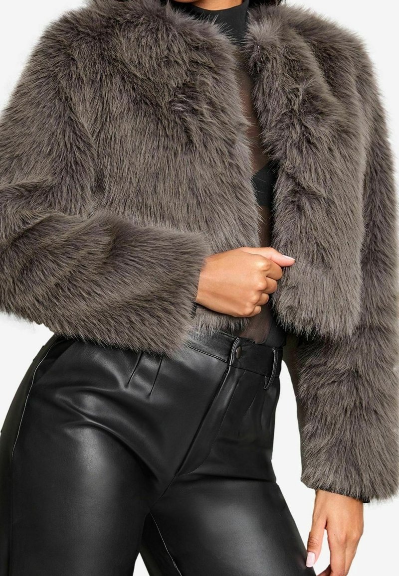 Femme portant une veste courte grise en fausse fourrure et un pantalon en cuir noir taille haute, une main reposant près de sa taille.