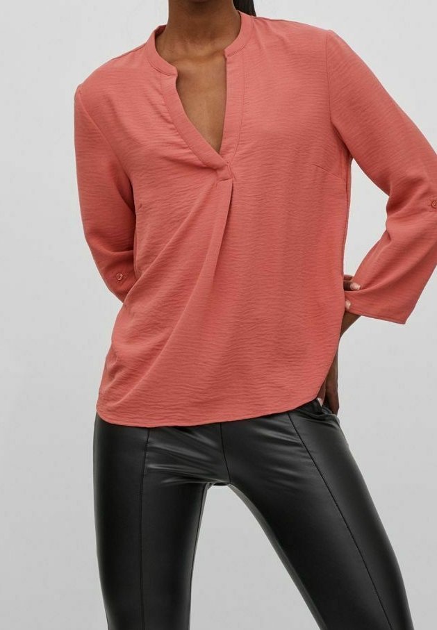 Personne portant un chemisier corail texturé à manches longues avec un col en V et un pantalon noir en similicuir, debout avec une main sur la hanche.
