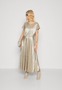 Robe maxi plissée dorée à manches courtes, texture brillante et taille élastique. Associée à des bottines beige à bout pointu et à une pochette en argent.