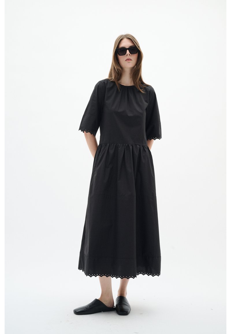 Femeie purtând o rochie midi neagră lejeră cu margini ondulate, pantofi slip-on negri și ochelari de soare negri pe un fundal alb.