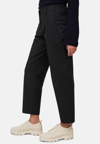 Persoon in zwarte cropped broek, marineblauwe trui en off-white chunky sneakers die zijwaarts staat op een witte achtergrond.