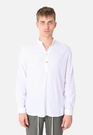 Giovane uomo con capelli corti che indossa una maglia henley bianca a maniche lunghe e pantaloni grigi su sfondo uniforme.