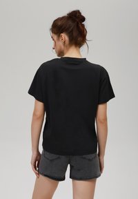 Camiseta negra de manga corta con un corte relajado, hecha de algodón, con cuello redondo y textura suave. Llevada con pantalones cortos de mezclilla grises.