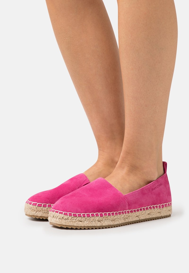 Pieds portant des espadrilles en daim rose vif avec des semelles en jute tressé et des semelles extérieures en caoutchouc sur un fond blanc.