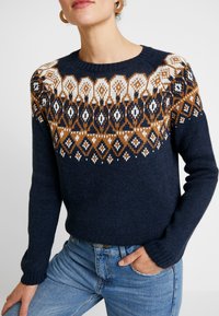Pull en tricot marine avec yoke à motifs en blanc et ambre. Col rond, manches longues et poignets côtelés. Associé à un jean bleu.