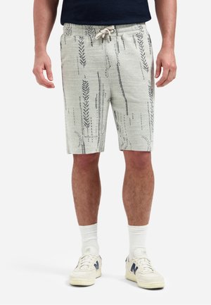 Man draagt beige shorts met trekkoord en zwarte abstracte lijnen, witte crew-sokken en witte New Balance sneakers, staand tegen een witte achtergrond.