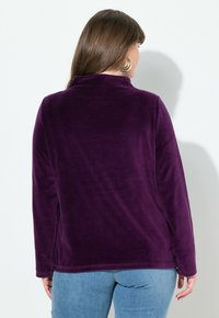 Lila Fleece-Oberteil mit langen Ärmeln, Kragen und seitlichen Schlitzen, strukturierte Oberfläche und lockere Passform, kombiniert mit hellblauen Jeans.