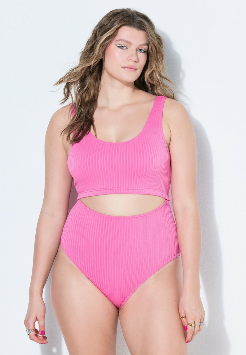 Donna con capelli mossi che indossa un costume da bagno a due pezzi rosa a coste con ritaglio sulla vita, in piedi davanti a uno sfondo bianco uniforme.