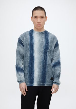 Jeune homme aux cheveux courts portant un pull tie-dye texturé bleu et gris et un pantalon noir, debout devant un fond blanc uni.