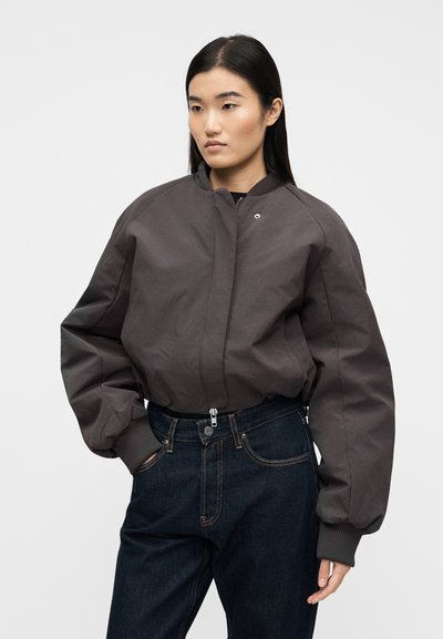 Γυναίκα με μακριά σκούρα μαλλιά φορώντας ένα γκρι σκούρο φαρδύ bomber μπουφάν και σκούρο μπλε ψηλόμεσα τζιν, με τα χέρια στις τσέπες.