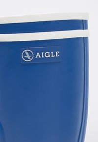 Bota de goma azul con ribete blanco en la parte superior, que presenta un parche de logo rectangular en relieve con el texto "AIGLE" y un emblema circular.