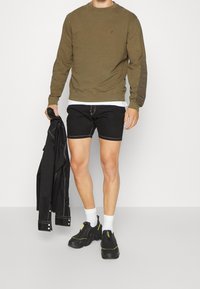 Olivgrön sweatshirt med rund halsringning, svarta denimshorts med vit sömnad, svart jacka och svarta skor med gula accenter.