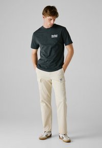 Mørk grå t-shirt med "RedBull Racing" logo, beige cargo bukser med lommer, og hvide sneakers med sorte og marineblå detaljer.
