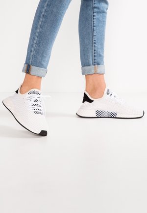 Osoba ubrana w podwinięte niebieskie dżinsy i białe siateczkowe trampki z czarnymi akcentami na białej podłodze.