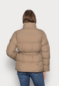 Beige pufferjakke med høj krave og belted talje. Har et quiltet design, lange ærmer og en glat, let tekstur.