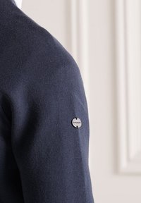 Maglione blu navy con una texture liscia, caratterizzato da un logo argentato sulla spalla sinistra. Design semplice, che enfatizza uno stile minimalista.