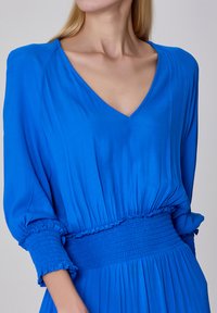 Robe bleue avec un décolleté en V, de longues manches bouffantes et une taille smockée. Tissu léger et fluide avec une texture subtile et des bords de manches volantés.