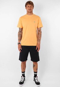 T-shirt orange à manches courtes avec un petit logo bleu, associé à un short noir et des baskets montantes noires, avec des accents blancs et des chaussettes.