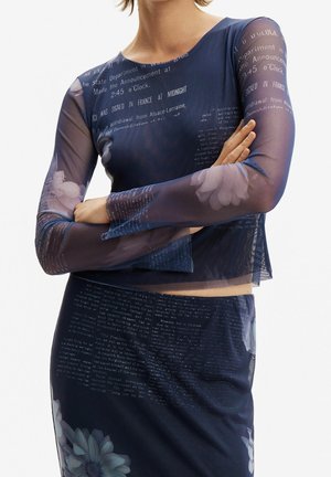 Kobieta ubrana w granatowy siateczkowy top z długim rękawem i dopasowaną spódnicę z białym kwiatowym i tekstowym nadrukiem, ze skrzyżowanymi na talii ramionami, na białym tle.