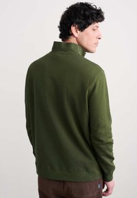 Hombre con cabello oscuro y rizado, vestido con un suéter de cremallera verde oliva y pantalones marrones, de perfil frente a un fondo claro y sencillo.