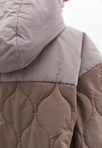 Padded bunda s designem béžovo-hnědého blokování barev, s prošívanou texturou na dolní části a hladkým materiálem na horní kapuci.