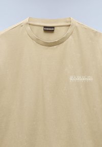 Camiseta de algodón beige con cuello redondo y mangas cortas. Presenta un logo impreso en blanco "NAPAPIJRI geographic" en el lado izquierdo del pecho.